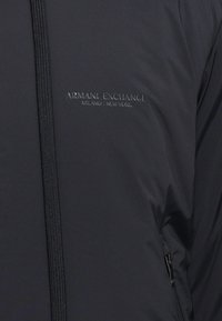 Sorte letvægtsjakke med lynlåslukning, standkrave og det prægede logo "ARMANI EXCHANGE" på venstre side.