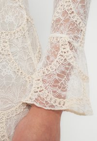 Lähikuva kädestä, joka on osittain peitetty läpikuultavalla kermaan värisellä pitsikankaalla, jossa on monimutkaisia kukka- ja silmukkareunuksia hihassa.