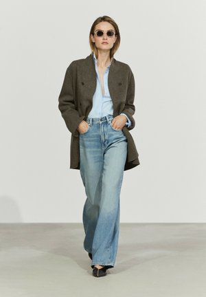 Vrouw met ronde zonnebril, bruine open jas, lichtblauw overhemd, jeans met wijde pijpen en zwarte puntige schoenen die op een lichtgrijze vloer loopt.