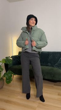 Chaqueta corta acolchada verde menta con cierre de cremallera, llevada sobre un suéter gris. Pantalones grises de pierna ancha y calcetines negros completan el conjunto.