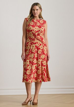 Czerwona i złota sukienka w kwiaty z detalem wiązanym w talii, krótkimi rękawami typu cap oraz zwiewną spódnicą, zestawiona z brązowymi butami na wysokim obcasie.