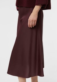 Frau in einem dunkelburgunderfarbenen Satin-Midirock und passendem gestrickten Pullover, mit der Hand entspannt an ihrer Seite vor einem schlichten hellen Hintergrund.