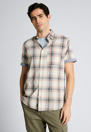 Młody mężczyzna w beżowo-szaro-czerwonej flanelowej koszuli na guziki z krótkim rękawem, noszący jasnoniebieski t-shirt i oliwkowe spodnie, stoi z jedną ręką w kieszeni.