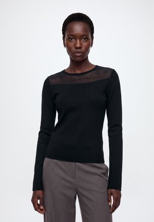 Femme aux cheveux courts et bouclés portant un haut noir à manches longues avec un panneau supérieur transparent et un pantalon gris taille haute, debout sur fond blanc.