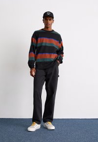 Black long-sleeve shirt with horizontal stripes in dark green, rust, and blue, paired with black loose-fit jeans and white sneakers with yellow laces.