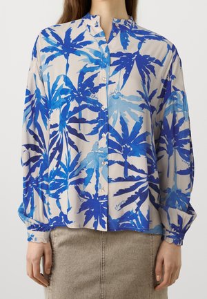 Kvinde iført en langærmet skjorte med knapper og et blåligt palmebladsmønster samt en beige nederdel.