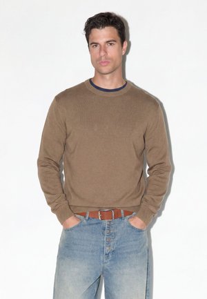 Mand med kort mørkt hår iført en brun sweater, blå jeans og brun bælte, står med hænderne i lommerne mod en hvid baggrund.
