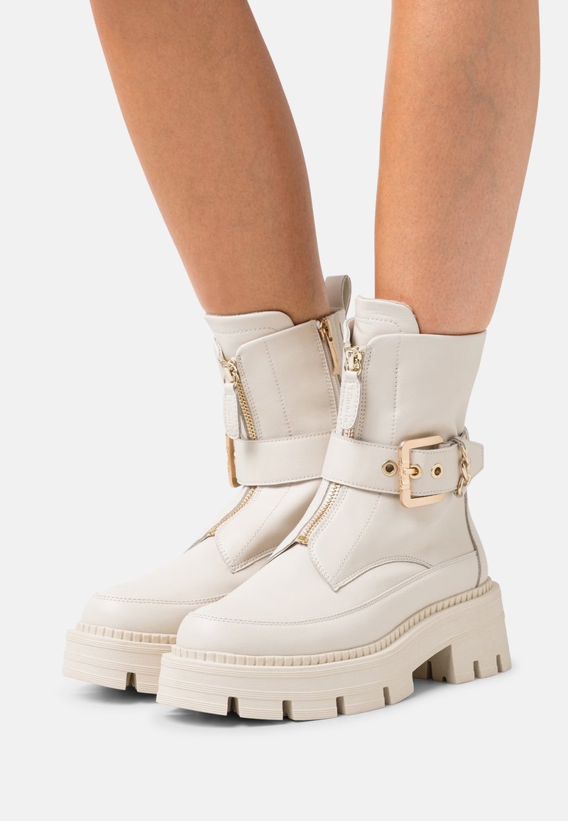 Botas de tobillo de cuero blanco con suela gruesa, que presentan dobles cremalleras y una correa decorativa con hebilla. Textura suave con un diseño moderno.