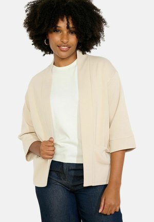 Frau mit lockigem Haar trägt eine beige offene Strickjacke, weißes Hemd und dunkelblaue Jeans, lächelt und posiert mit der Hand auf der Strickjacke.