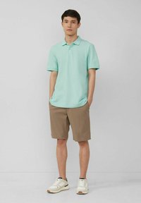 Mintgrünes Poloshirt und khaki Shorts, kombiniert mit weißen Turnschuhen. Das Shirt hat einen klassischen Kragen und kurze Ärmel.