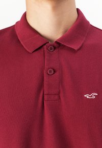 Vínový polo triko s límečkem, obsahující dvě knoflíkové zapínání a malé bílé vyšívané logo na levé straně. Texturovaná tkanina.