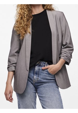 Vrouw met krullend haar die een grijze blazer draagt over een zwart shirt en blauwe spijkerbroek, met één hand in haar zak en de andere ontspannen langs haar zij.