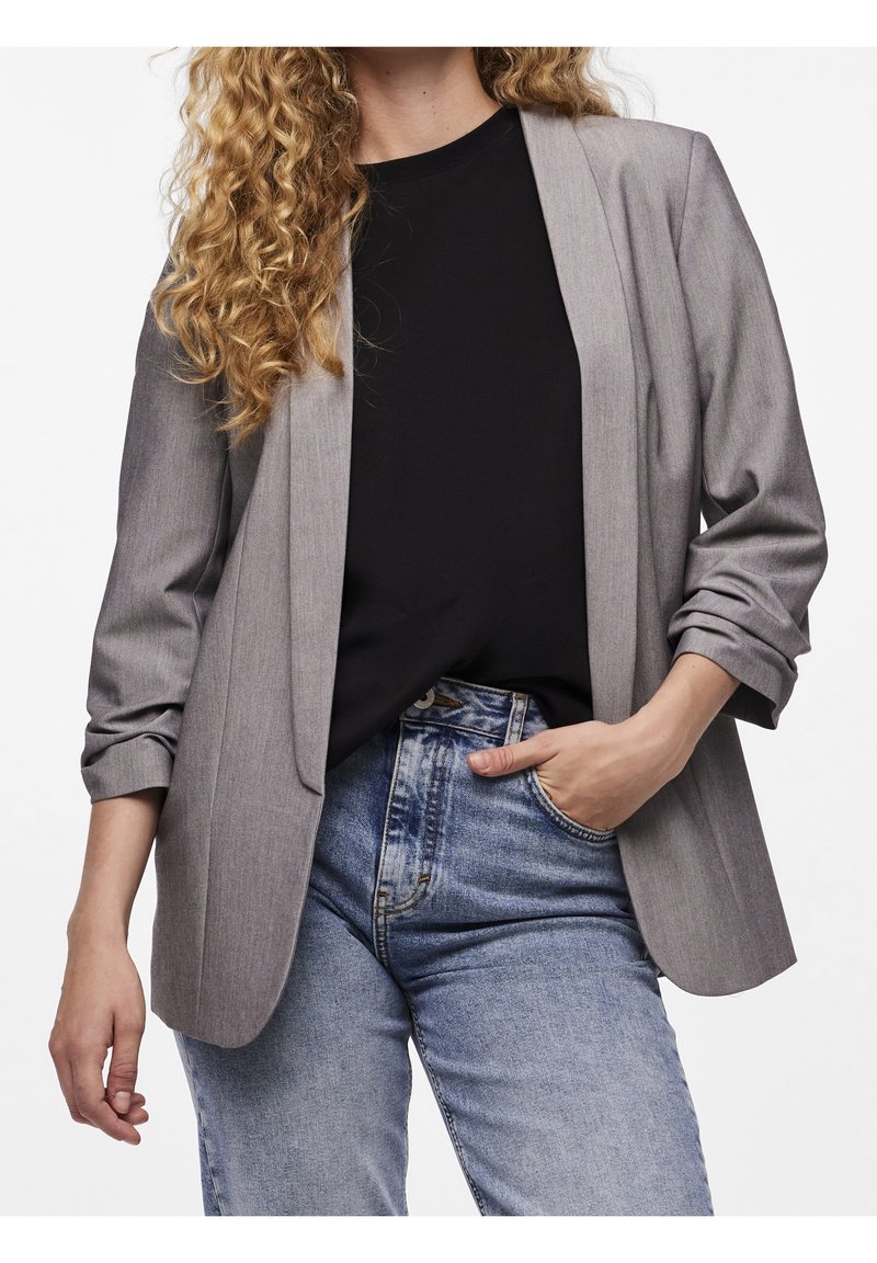 Femme aux cheveux bouclés portant un blazer gris sur une chemise noire et un jean bleu, une main dans la poche et l'autre détendue sur le côté.