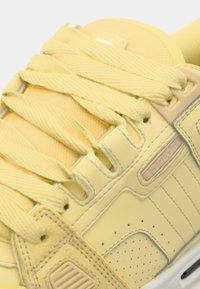 Baskets jaunes en tissu texturé, avec des lacets plats et des accents en œillets. Présentent de subtils perforations et un renfort au niveau de la pointe.