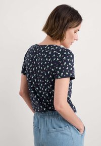 Jūros laivyno trumpomis rankovėmis marškinėliai su žydinčių gėlių raštu žaliai ir rožine spalvomis. Minkšta medžiaga, apvalus iškirpimas ir laisvas siluetas, derinami su šviesiais džinsiniais kelnais.