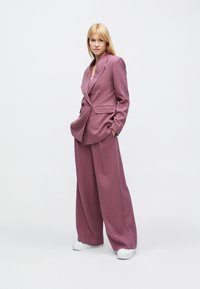 Maßgeschneiderter zweiteiliger Anzug aus weichem malvenfarbenem Stoff, bestehend aus einem zweireihigen Blazer mit Fronttaschen und weitem Hosenbein, kombiniert mit weißen Schuhen.