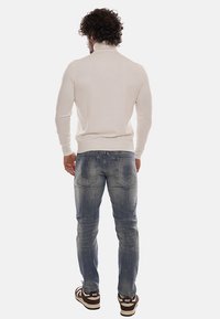Pullover a collo alto color crema, design aderente, abbinato a jeans chiari strappati con dettagli del logo, e sneakers marroni con accenti bianchi.