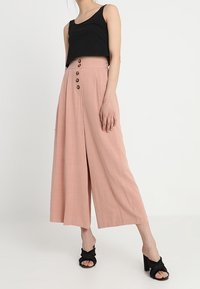 Pink wide-leg trousers made of lightweight fabric, featuring a high waist and five buttons, paired with a black sleeveless top and black sandals.