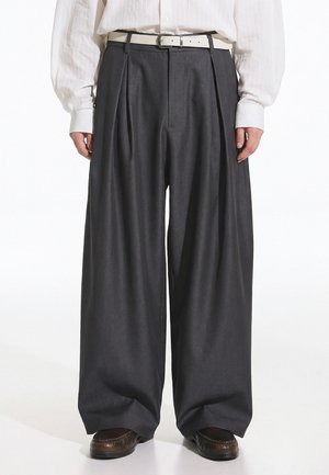 Személy sötét, bő szárú nadrágot, fehér övet, fehér hosszú ujjú inget és barna mokaszint visel, fehér háttér előtt áll.