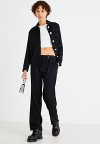 Chaqueta negra texturizada con botones blancos, combinada con pantalones negros de tiro alto. La modelo lleva botas gruesas de plataforma negras y sostiene un pequeño bolso.