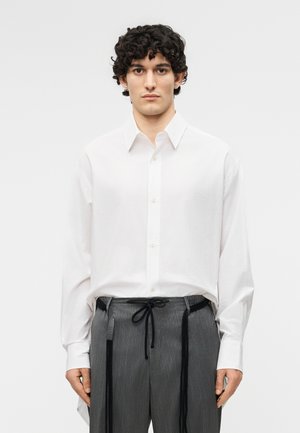 Νεαρός με σγουρά μαλλιά, φορώντας λευκό πουκάμισο με κουμπιά και γκρι παντελόνι με λεπτές ρίγες και μαύρα κορδόνια, στέκεται μπροστά σε μια απλή φόντο.