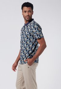 Kortärmad polo t-shirt med marinblå bakgrund, prydd av ett färgglatt palmbladsmönster. Kombineras med ljus beige byxor, med avslappnade designelement.