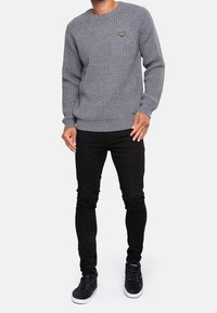 Grijze gebreide trui met een textuurpatroon, geribde manchetten en zoom, gecombineerd met zwarte slim-fit jeans en zwarte sneakers. Gele logo-accent op de borst.