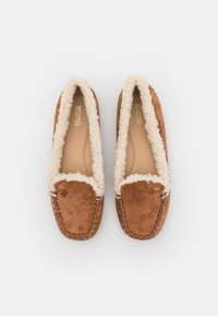 Brune mockasinslipper med en myk kremfarget lammeskinnsfôr, mokkasinstil søm og en gulllogo-akseent på siden.