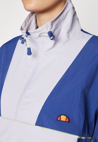 Lagani ljubičasti i plavi jakni s visokim ovratnikom, podešivim trakama i izvezenim logotipom u narančastoj i žutoj boji.