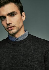 Pull en laine noir à col rond, superposé à une chemise bleu clair à boutons, avec un col contrastant. Textures subtiles visibles.