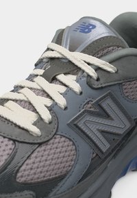 Gros plan sur une basket grise et lavande avec des lacets blancs, des panneaux en mesh et un logo "N" prominent sur le côté.