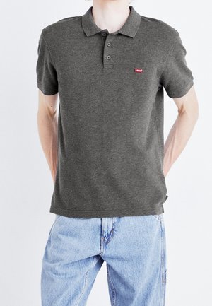 Personne portant un polo Levi's gris foncé et un jean bleu clair, debout les mains derrière le dos sur un fond uni.