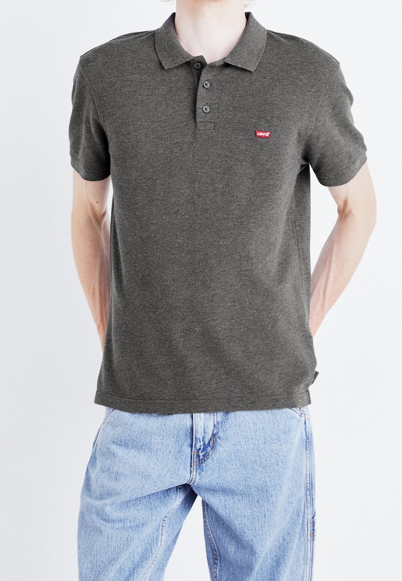 Personne portant un polo Levi's gris foncé et un jean bleu clair, debout les mains derrière le dos sur un fond uni.