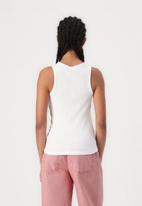 Weißes, geripptes Tanktop mit V-Ausschnitt und ärmellosem Design, kombiniert mit rot gestreiften Hosen. Glatte Textur, figurbetonte Silhouette.