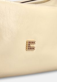 Crèmekleurige leren tas met een gestructureerde afwerking, voorzien van een opvallende letter "E" in metalen zeshoekige studs in goud en zilver.