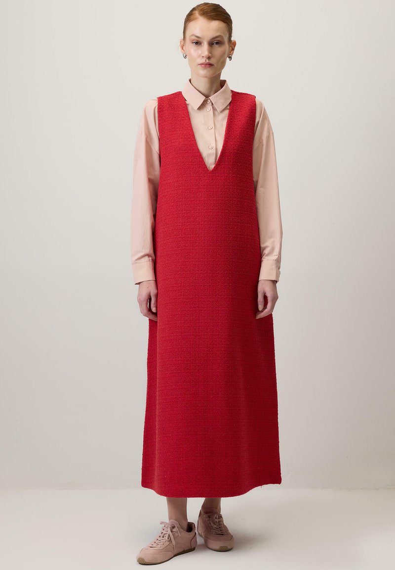 Červené bezrukávové šaty s hlbokým V-výstrihom, štruktúrovaná tkanina a dlhý, rovný silueta; nosené cez svetloružovú košeľu a v páre s béžovými topánkami.