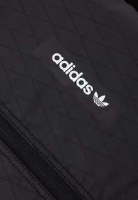 Černá prošívaná tkanina s diagonálním vzorem, se bílým logem a emblémem Adidas. Obsahuje černý detail s zipem.