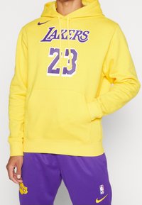 Man i en gul Lakers-huvtröja med nummer 23 och lila Lakers-baskettights med NBA- och Nike-logotyper, hand i huvrtröjans ficka.