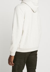 Persona vestida con una sudadera beige clara y pantalones cargo verde oscuro, de pie con la espalda y el lado izquierdo parcialmente visibles contra un fondo liso.