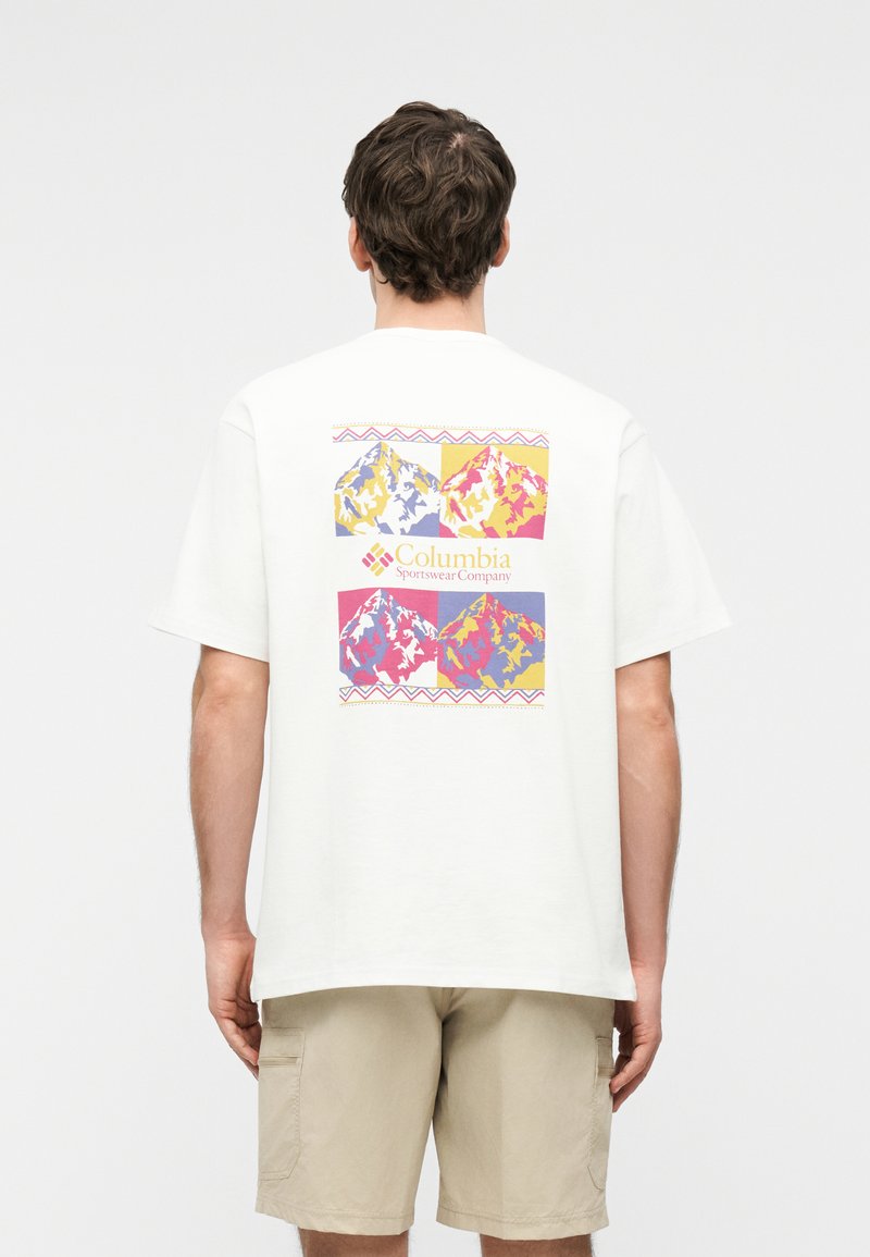 Άνδρας που φοράει λευκό Τ-shirt με πολύχρωμο γραφικό βουνό και το κείμενο «Columbia Sportswear Company» στην πλάτη, συνδυασμένο με μπεζ σορτς.
