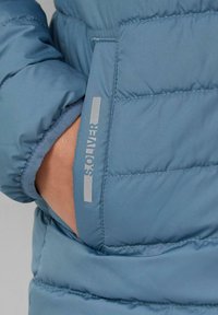 Hellblaue Steppjacke mit gestepptem Design, ausgestattet mit einer seitlichen Tasche. Enthält ein silbernes "Solver"-Logo am Ärmel.