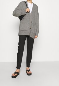 Grå stickad cardigan med en lång, lös passform, utrustad med två framfickor och stora knappar, kombinerad med svarta slim-fit byxor och svarta sandaler.