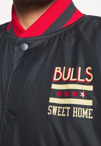 Sort bomberjakke med røde detaljer. Har et grafisk design med teksten "BULLS SWEET HOME" og stjerner. Mat finish og trykknaplukning.