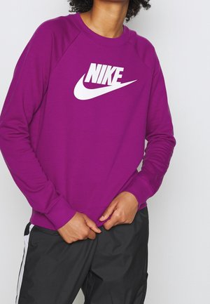 Persoon draagt een felpaarse Nike-trui met wit logo, gecombineerd met een zwarte broek met witte strepen, handen dicht bij de taille samengevouwen.