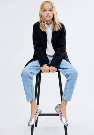 Silberne Ballerinas, hellblaue Jeans, cremefarbener Pullover und ein schwarzer Mantel. Modell sitzt auf einem Hocker vor einem schlichten Hintergrund.