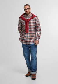 Mann mit Brille, der ein rot kariertes Hemd, einen roten Pullover um die Schultern gebunden, blaue Jeans und braune Wanderschuhe trägt, vor einem weißen Hintergrund steht.