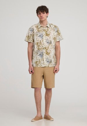Camicia a maniche corte, di colore chiaro, con bottoni, caratterizzata da un motivo di foglie verdi e blu, abbinata a pantaloni corti beige e scarpe slip-on color cammello.