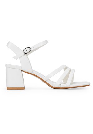 Sandalias de tacón blanco con tacón bloque, que presentan un diseño con tiras y hebilla ajustable en el tobillo. Textura de cuero suave con una suela color marrón.