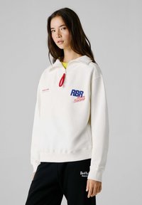 Νεαρή γυναίκα που φοράει λευκό φούτερ με μισή ζώνη και το λογότυπο "RBR Team Racing" και μαύρο παντελόνι με το λογότυπο "Red Bull Racing".