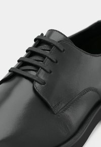 Μαύρα δερμάτινα παπούτσια με λείο, γυαλιστερό επιφάνειας, στρογγυλή μύτη και παραδοσιακό σύστημα κορδονιών. Διαθέτει λεπτομέρεια από διακριτική ραφή.